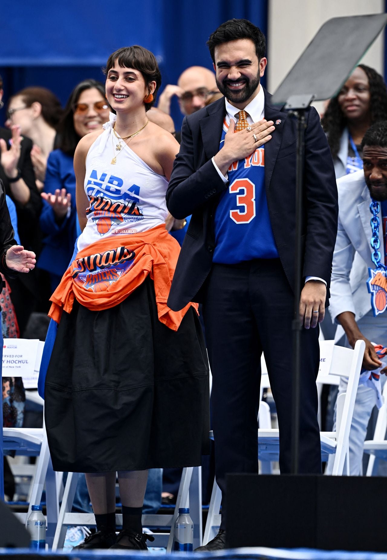 Two people on a stage with a crowd in the background, one wearing an NBA jersey.