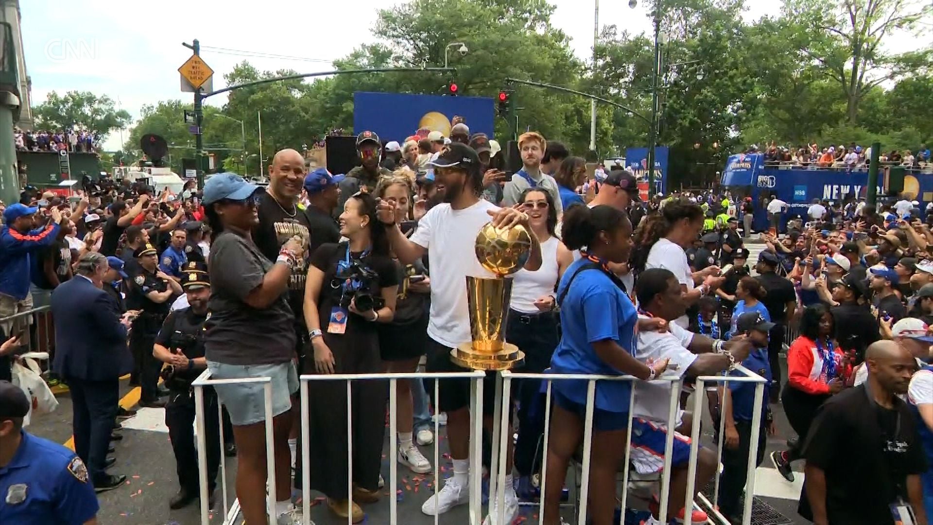 Person holding a trophy in front of a crowd on a street, with trees and buildings in the background