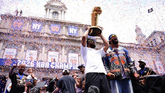 Two men celebrating with a trophy in front of a large crowd and building with 'NY' on it.